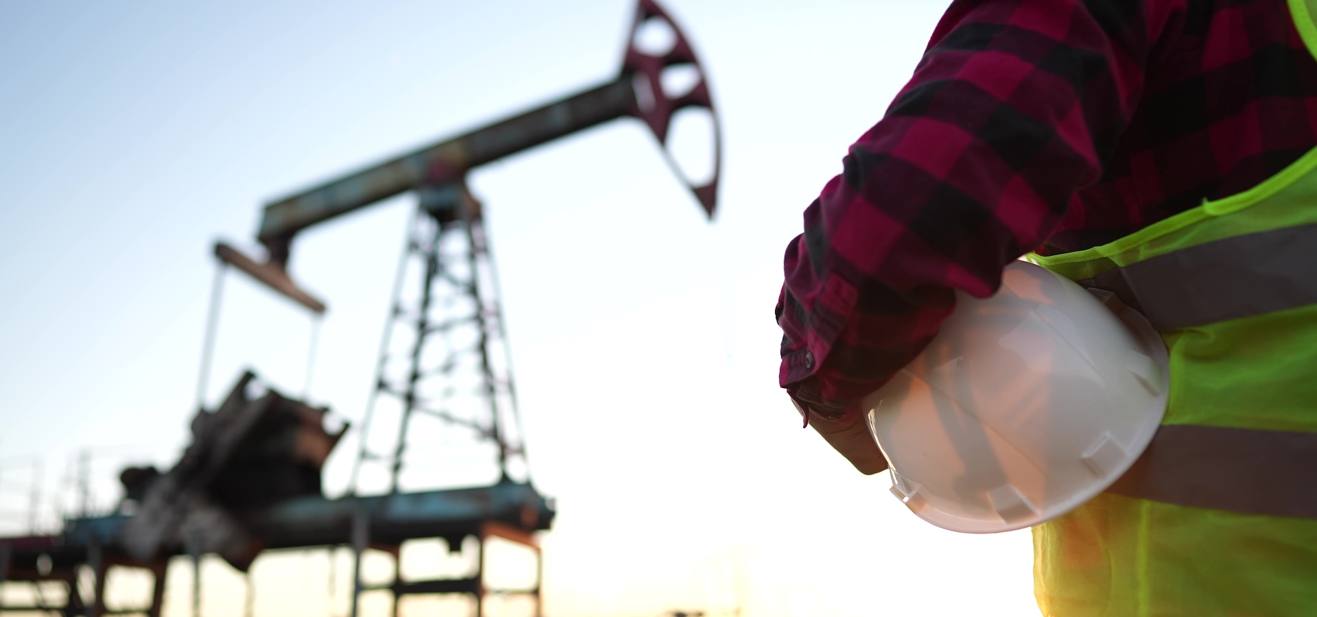 a worker holding protective hard hat at sunset in the background an oil pump